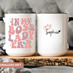 May include: Two white ceramic mugs with black handles. One mug has the text "In My Boss Lady Era" in pink and blue lettering with a crown and heart design. The other mug has the text "Sophia" with a crown design.