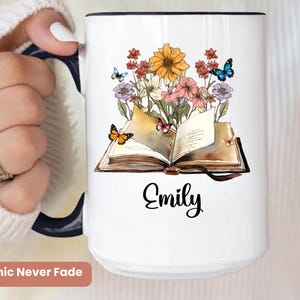 May include: White ceramic mug with a black handle and rim. The mug features a floral and butterfly design with an open book and the name "Emily" in black script. The words "Graphic Never Fade" are at the bottom.