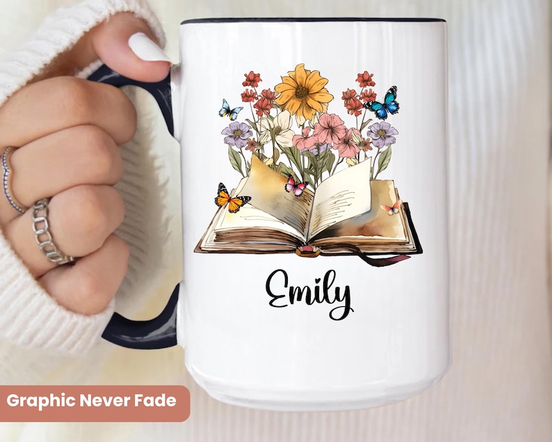 May include: White ceramic mug with a black handle and rim. The mug features a floral and butterfly design with an open book and the name "Emily" in black script. The words "Graphic Never Fade" are at the bottom.