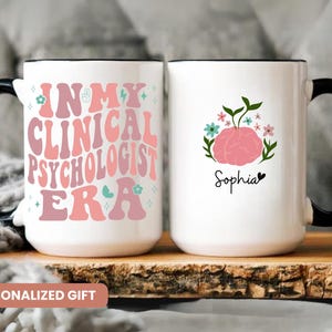 May include: Two white ceramic mugs with black handles. One mug has a pink and white design with the text "In My Clinical Psychologist Era". The other mug has a pink brain with flowers and the name "Sophia".