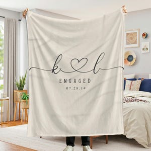 May include: A white throw blanket with the initials "K" and "L" in a cursive font, connected by a heart. The text "ENGAGED" and the date "07.28.14" are printed below the initials.