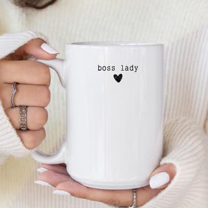 Pu&ograve; includere: Una tazza di ceramica bianca con la scritta "boss lady" in nero, accompagnata da un piccolo cuore nero. La tazza ha un manico grande ed &egrave; tenuta da una persona che indossa un maglione bianco.