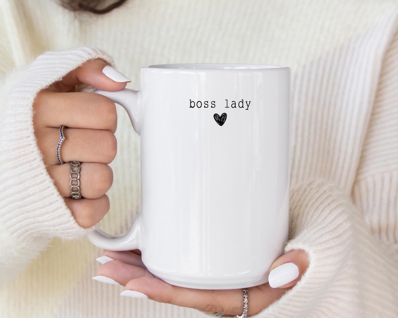 Pu&ograve; includere: Una tazza di ceramica bianca con la scritta "boss lady" in nero, accompagnata da un piccolo cuore nero. La tazza ha un manico grande ed &egrave; tenuta da una persona che indossa un maglione bianco.