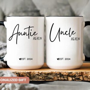 May include: Two white ceramic mugs with black handles. The mug on the left says "Auntie Again" with a heart and "EST. 2024" below. The mug on the right says "Uncle Again" with a heart and "EST. 2024" below.