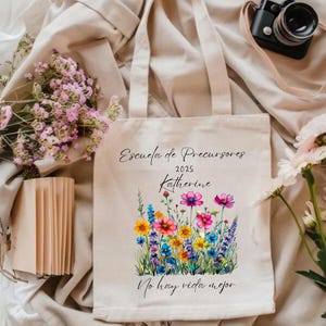 May include: A beige tote bag with a floral design featuring colorful wildflowers. The bag has the text "Escuela de Precursores 2025 Katherine" and "No hay vida mejor" printed on it. The bag has two handles and is made of a canvas-like material.