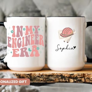 May include: Two white ceramic mugs with black handles. One mug has the text "In My Engineer Era" in pink and blue letters with small flowers and lightning bolts. The other mug has a pink hard hat and the name "Sophia" in black script.