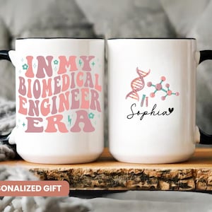 May include: Two white ceramic mugs with black handles. The mug on the left has pink and blue text that reads "In My Biomedical Engineer Era". The mug on the right has a pink and blue DNA strand and molecule graphic with the name "Sophia" in black script.