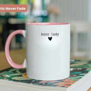 Pu&ograve; includere: Tazza in ceramica bianca con interno e manico rosa. La tazza presenta la scritta "boss lady" sopra un piccolo cuore nero. Le parole "Graphic Never Fade" sono in un rettangolo rosa in alto a sinistra.