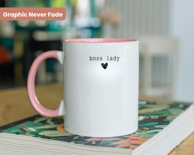 Pu&ograve; includere: Tazza in ceramica bianca con interno e manico rosa. La tazza presenta la scritta "boss lady" sopra un piccolo cuore nero. Le parole "Graphic Never Fade" sono in un rettangolo rosa in alto a sinistra.