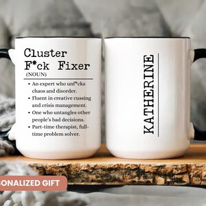 May include: White ceramic mug with black handle and rim. The mug features the text "Cluster F*ck Fixer" with a definition, and the name "KATHERINE" on the other side. The mug is a personalized gift.