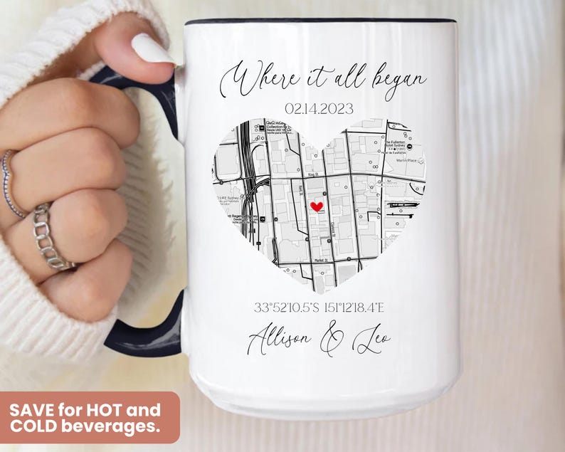 May include: White ceramic mug with a black rim and handle. The mug features a black and white map of a city in the shape of a heart with a red dot in the center. The text "Where it all began" is printed above the map, followed by the date "02.14.2023". Below the map, the coordinates "33&deg;52'10.5'S 151&deg;12'18.4'E" and the names "Allison & Lee" are printed.