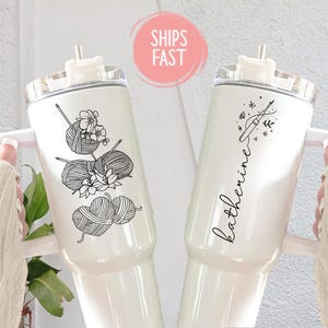 May include: Two white insulated tumblers with handles. One features a black illustration of yarn and flowers, the other has a knitting needle design and the name "Katherine" in script. Both have a straw and a clear lid.