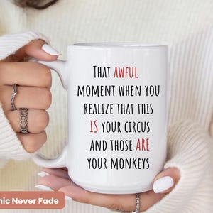 Op de afbeelding: Witte keramische mok met de tekst "THAT AWFUL MOMENT WHEN YOU REALIZE THAT THIS IS YOUR CIRCUS AND THOSE ARE YOUR MONKEYS" in zwarte en rode letters. De mok wordt vastgehouden door een persoon die een witte trui draagt. De woorden "Graphic Never Fade" staan onderaan.