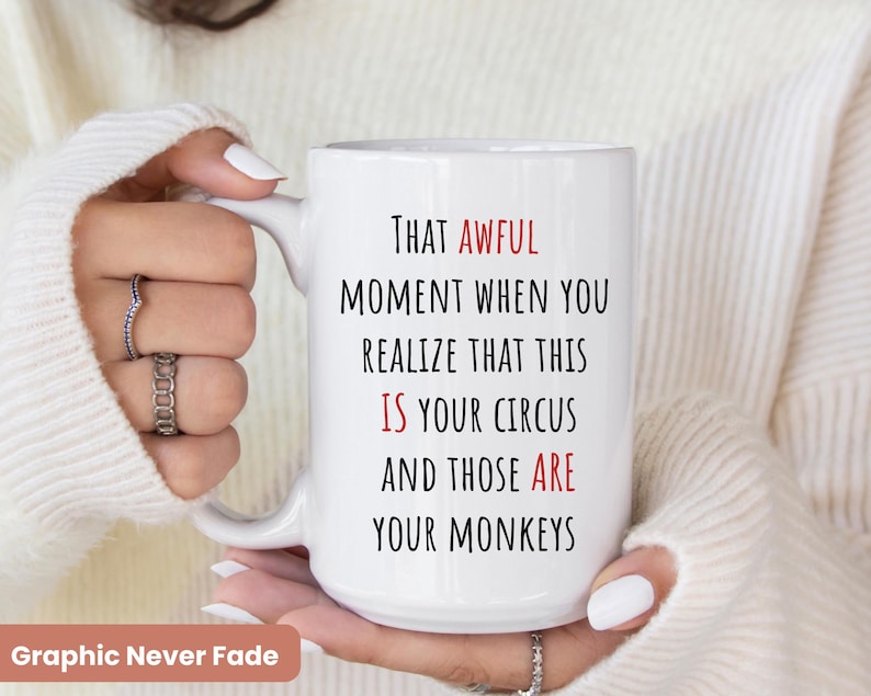 Op de afbeelding: Witte keramische mok met de tekst "THAT AWFUL MOMENT WHEN YOU REALIZE THAT THIS IS YOUR CIRCUS AND THOSE ARE YOUR MONKEYS" in zwarte en rode letters. De mok wordt vastgehouden door een persoon die een witte trui draagt. De woorden "Graphic Never Fade" staan onderaan.