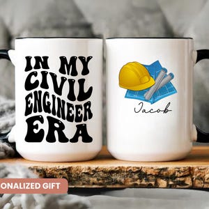 May include: Two white ceramic mugs with black handles. One mug has the text "In My Civil Engineer Era" in black. The other mug has a blue and white drawing of a hard hat and blueprints with the name "Jacob" in black.