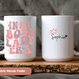 May include: Two white ceramic mugs with a pink and green design. One mug says "In My Boss Lady Era" and the other mug says "Sophia" with a pink crown above the name.