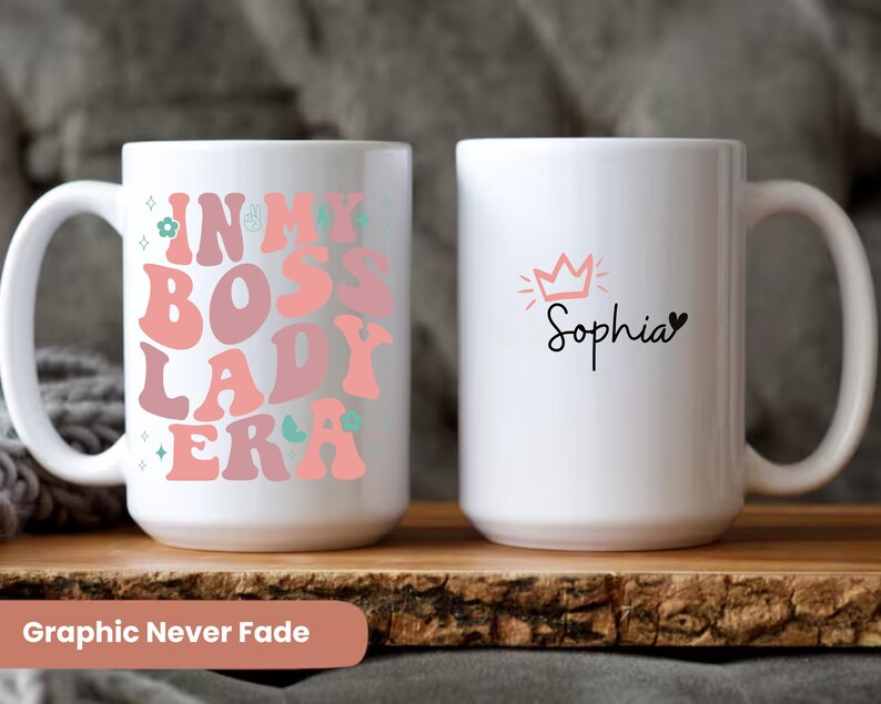 May include: Two white ceramic mugs with a pink and green design. One mug says "In My Boss Lady Era" and the other mug says "Sophia" with a pink crown above the name.