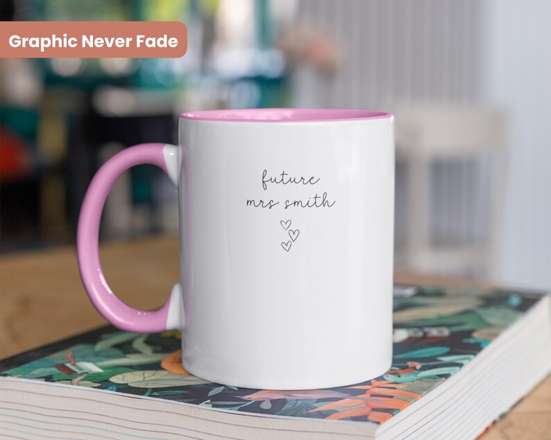 Pu&ograve; includere: Tazza in ceramica bianca con bordo e manico rosa. La tazza ha il testo "future mrs smith" scritto su di essa in corsivo nero con tre piccoli cuori sotto il testo.