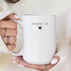 May include: White ceramic mug with the text "chapter 60" and a black heart. The mug is held by a person wearing a white sweater. The mug is a standard size for coffee or tea.