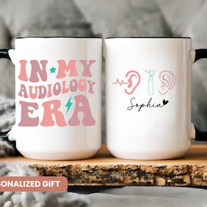 May include: Two white ceramic mugs with black handles. The mug on the left has the text "In My Audiology Era" in pink and blue lettering with a star and lightning bolt. The mug on the right has a heart, the text "Sophia", and a stylized image of an ear, an otoscope, and a heartbeat. Both mugs are on a brown wooden surface.