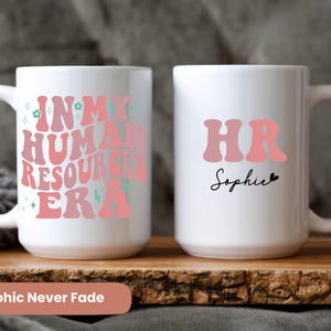 May include: Two white ceramic mugs with pink and green text. One mug says "In My Human Resources Era" and the other mug says "HR Sophie".