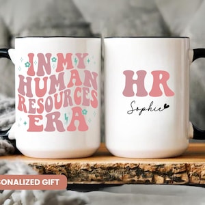 May include: Two white ceramic mugs with black handles. The mug on the left has the text "In My Human Resources Era" in pink and blue lettering with a lightning bolt and flower accents. The mug on the right has the text "HR Sophie" in pink lettering with a small heart.