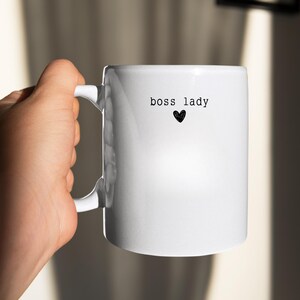 Pu&ograve; includere: Una tazza di ceramica bianca con le parole "boss lady" stampate in nero, insieme a un piccolo cuore nero. La tazza ha un manico curvo ed &egrave; tenuta da una mano.