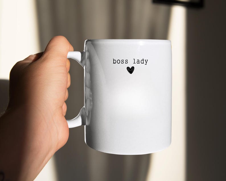 Pu&ograve; includere: Una tazza di ceramica bianca con le parole "boss lady" stampate in nero, insieme a un piccolo cuore nero. La tazza ha un manico curvo ed &egrave; tenuta da una mano.