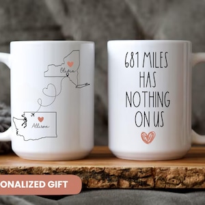 May include: Two white ceramic mugs with black text and pink hearts. The mug on the left has a map of the United States with the states of New York and Washington highlighted. The mug on the right has the text "681 miles has nothing on us".