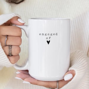 May include: White ceramic mug with the text "engaged af" in black, accompanied by a black heart symbol. The mug has a large handle and is held by a person wearing a white sweater.