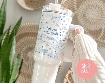 May include: White travel tumbler with blue illustrations and text. The tumbler features a handle and a lid with a straw. The text reads "future mrs. Smith" and "02.03.2026" with wedding-themed graphics. The tumbler is held by a person wearing a white sweater.