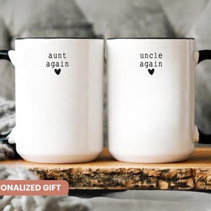 May include: Two white ceramic mugs with black handles and rims. One mug reads "aunt again" and the other reads "uncle again", each with a small black heart. The mugs are on a wooden surface.