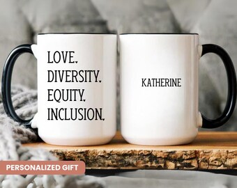 May include: Two white ceramic mugs with black handles. One mug has the text "LOVE. DIVERSITY. EQUITY. INCLUSION." printed on it. The other mug has the name "KATHERINE" printed on it. The mugs are sitting on a wooden surface.