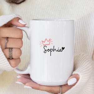 May include: White ceramic mug with a pink crown and the name "Sophia" written in black cursive with a small black heart next to it.