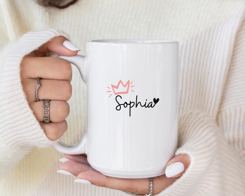 May include: White ceramic mug with a pink crown and the name "Sophia" written in black cursive with a small black heart next to it.