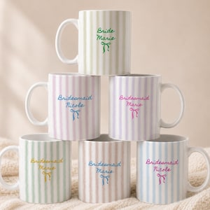 May include: A stack of white ceramic mugs with vertical striped patterns in pastel colors. Each mug features a name and a bow graphic in a coordinating color. The mugs are for a bridal party.
