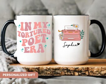 May include: Two white ceramic mugs with black handles. One mug has the text "In My Tortured Poet Era" in pink and blue lettering. The other mug has a pink typewriter with flowers and the name "Sophia" written below it.
