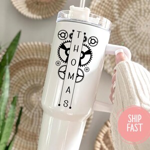 May include: White tumbler with a handle, featuring the name "THOMAS" in black, stylized with gears and mechanical elements. The tumbler has a clear lid and a straw. The background includes woven wall decor and greenery.
