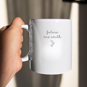 Pu&ograve; includere: Tazza in ceramica bianca con il testo "future mrs. smith" e tre piccoli cuori.