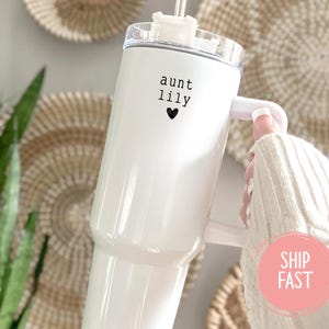 May include: A white insulated tumbler with a handle, featuring the text "aunt lily" and a black heart. The tumbler is held by a person wearing a white sweater. The background includes woven wall decor and green plants.