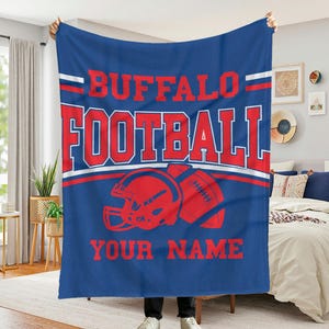 May include: A blue blanket with the words "BUFFALO FOOTBALL" in red block letters, along with a red football helmet and football graphic. The words "YOUR NAME" are printed below the graphic. White stripes accent the top and middle of the blanket.