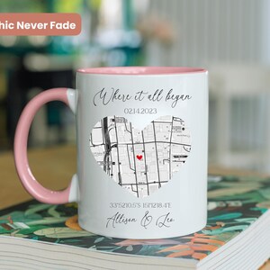 May include: White ceramic mug with a pink rim and handle. The mug features a black and white map of a city in the shape of a heart with a red dot in the center. The text "Where it all began" is above the map, and the date "02142023" is below the map. The coordinates "33.5210.5'S 151.218.4'E" and the names "Allison & Leo" are below the coordinates.