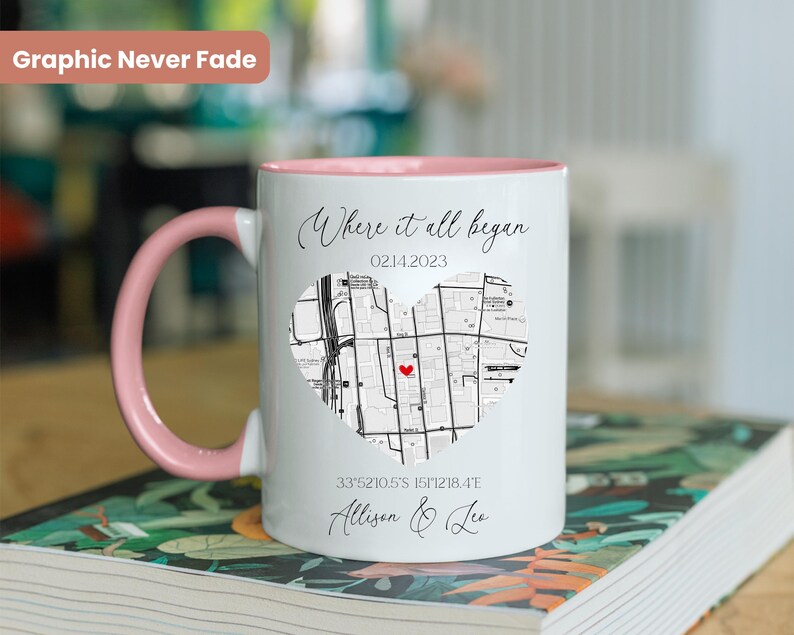 May include: White ceramic mug with a pink rim and handle. The mug features a black and white map of a city in the shape of a heart with a red dot in the center. The text "Where it all began" is above the map, and the date "02142023" is below the map. The coordinates "33.5210.5'S 151.218.4'E" and the names "Allison & Leo" are below the coordinates.