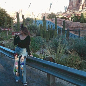 Könnte beinhalten: Eine Frau trägt ein schwarzes Oberteil und eine blaue Jeans mit einem Mickey Mouse und Freunde-Druck. Sie steht vor einem Metallgeländer mit einer Wüstenlandschaft im Hintergrund.