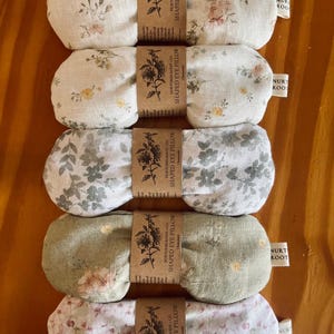 May include: Five eye pillows with floral patterns in white, green, and pink. Each pillow is wrapped with a brown paper band that reads "SHAPED EYE PILLOW". The pillows are arranged on a wooden surface. The pillows are approximately 20 cm long.