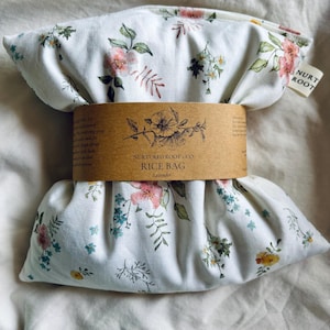May include: A rectangular rice bag made of white fabric with a floral pattern featuring pink, blue, and yellow flowers. A brown paper band wraps around the center, printed with the text Nurtured Root + Co. Rice Bag Lavender and a floral illustration.
