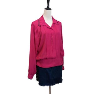 May include: A long-sleeved, fuchsia blouse with a collared neckline and black trim. The blouse features vertical pleats down the front and a gathered waist. It is paired with a dark denim skirt.