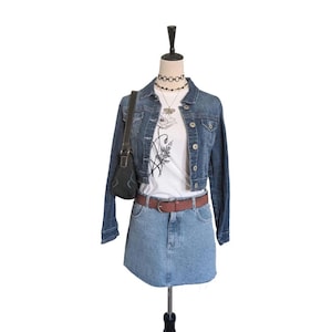 May include: A denim jacket, white graphic tee, and denim skirt outfit. The jacket is a dark wash, cropped style. The skirt is a light wash, mini skirt. A brown belt and black purse complete the look. A black choker and necklace are also included.