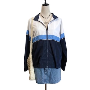 Puede incluir: Chaqueta con bloques de color azul marino, blanco y azul claro, con cremallera completa y cuello alzado. La chaqueta se combina con una falda vaquera y un bolso tote de ganchillo color crema. Maniquí.