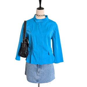 May include: A mannequin displays a turquoise blue jacket with a high collar and three-quarter sleeves, paired with a denim skirt. A black handbag with a decorative pattern hangs from the mannequin's shoulder. A black choker necklace is also visible.